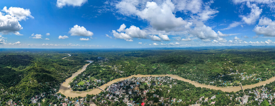 Aerial view of the Sangu River winding through the lush green hills and the town of Bandarban under a bright blue sky with scattered clouds in Bandarban, Chittagong Division, Bangladesh.