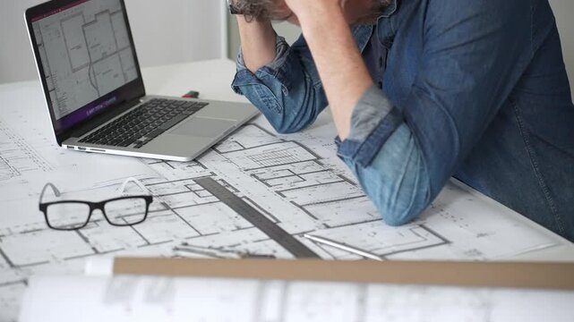 Architect sketching building plans on paper using a ruler and pen while working digitally on a laptop with a blueprint software displaying a floor plan