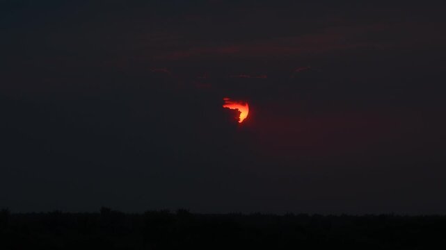 A time‑lapse shot: a red sun, partially obscured by clouds, is sinking below the horizon, creating a striking contrast with the dark sky and the silhouettes of the trees.