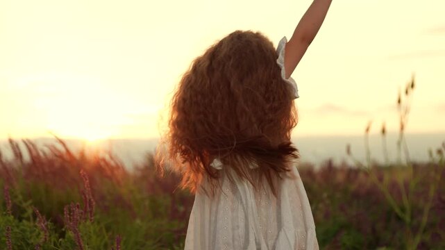 Funny little girl dances raising hands in wild field at summer sunset backside view. Little girl dances and has fun in rural field on summer weekend. Happy little girl dances in park field at sunset
