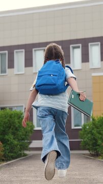 child kid girl runs book hands class, child kid girl run school backpack against background school, little girl daughter runs across school yard education building, energetic child happy schoolboy