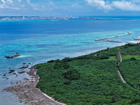 Aerial view of Taketomi Island featuring lush green vegetation, rocky shoreline, turquoise coral reef waters, and a distant coastal town in Taketomi, Okinawa, Japan.