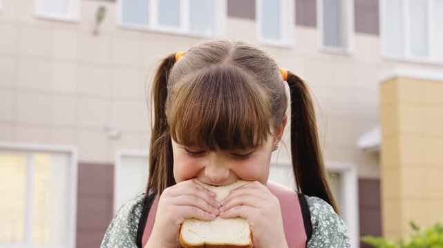 Little girl with pigtails munching sandwich, Kid takes a bite out of school lunch, Childhood innocence at lunchtime, Student with backpack and big appetite, Girl savoring tasty sandwich after school