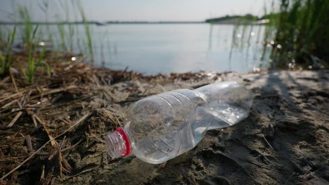 A crushed plastic bottle lies discarded on a sandy lake shoreline. Wild grass and calm water surround the single use waste under bright daylight, highlighting environmental pollution.