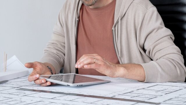 Architect wearing glasses using a digital tablet at office desk, reviewing blueprints and building model, focusing on modern residential design and innovation in architecture