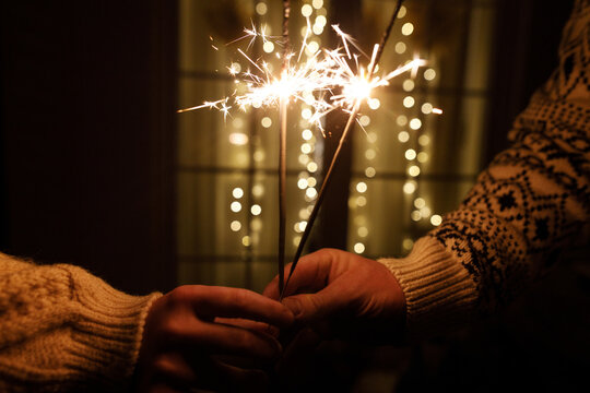 Couple Hands Holding Burning Sparklers in Warm Cozy Sweaters with Golden Christmas Lights Bokeh Background on New Year Eve. Happy new year, atmospheric eve