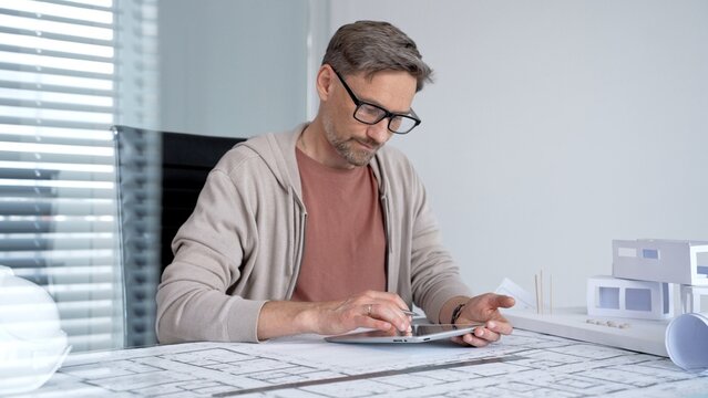 Architect wearing glasses using a digital tablet at office desk, reviewing blueprints and building model, focusing on modern residential design and innovation in architecture