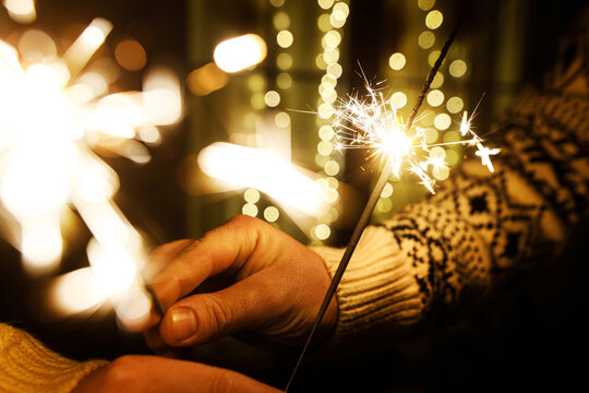 Close-up of hands holding glowing sparkler with bright light bokeh, cozy sweater. Holiday warmth, celebration and magic of New Year and  Christmas.
