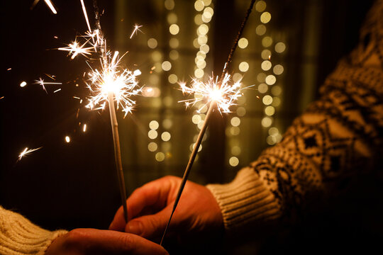 Hands holding burning sparklers against golden Christmas bokeh lights background. Cozy holiday mood, celebration of New Year, intimacy and warmth.