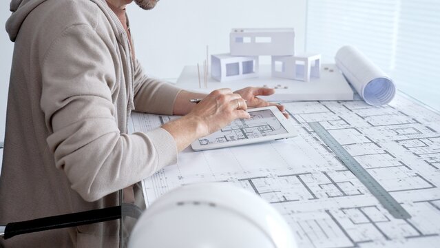 Architect working on a new building design using a tablet with digital blueprint plans. Also showing paper blueprints. A ruler. And a scale model of a house on the desk