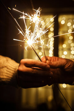 Vertical Close Up of Hands Holding Burning fireworks with Warm Festive Garland Lights. Christmas wonder and winter holidays. Burning Sparklers on New Year Eve