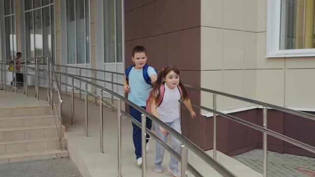 School kids using accessibility ramp, Children hold onto metal rails, The surface is smooth and flat, The background shows big windows, They wear comfortable clothes, Their backpacks are colorful, The