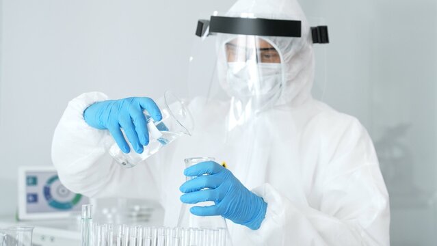 Scientist wearing a hazmat suit. Face shield. And protective gloves pouring liquid from a beaker into an erlenmeyer flask