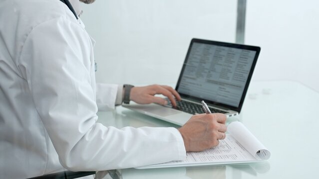 Healthcare professional wearing a white lab coat and stethoscope, sitting at a desk and writing patient notes on a clipboard and using a laptop in a modern clinic office