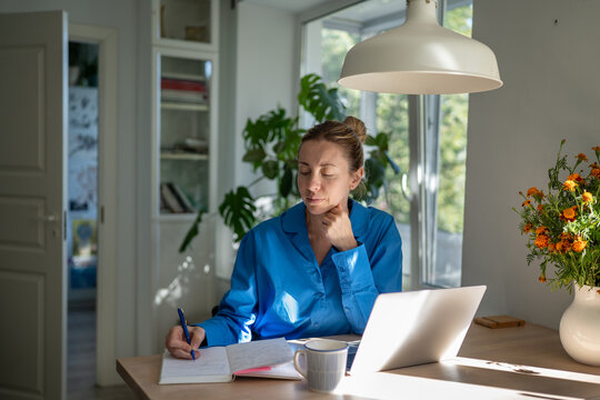 Focused woman working from home sitting at table with laptop, writing plans and ideas in notebook, thoughtful calm female remote worker taking, notes in diary at desk with computer in kitchen.