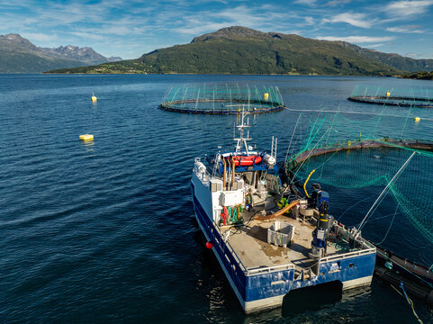 Aerial view of a salmon farm in Gratangsfjorden with a work boat moored alongside circular net pens in the deep blue water surrounded by mountains in Gratangsbotn, Troms, Norway.