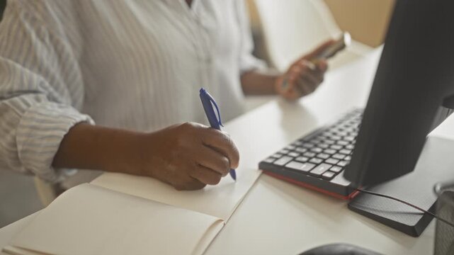 Woman writing with blue pen and holding smartphone at office desk near computer keyboard; concentration productivity.