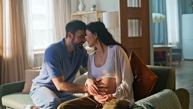 Married couple bonding pregnant belly home sofa. Man touching wife baby bump.