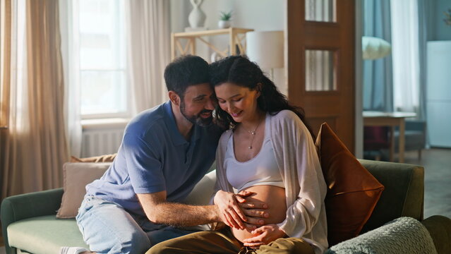 Loving couple touching pregnant belly home sofa. Man wife tender bonding.
