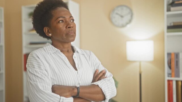 Woman with arms crossed and watch, standing in an interior room with bookshelf and clock inside a building; determination reflection.