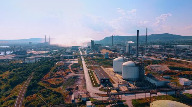Heavy manufacturing plant with detailed metal pipelines running across the structures. Plumes of white smoke rise from the smokestacks in the backdrop. Hills in the backdrop.