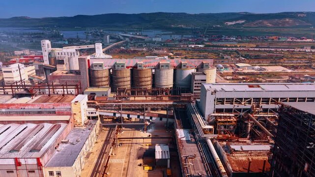Sprawling industrial complex featuring massive concrete silos, intricate piping networks, and large factory buildings. Coastal port area with cranes and green hills in the backdrop.