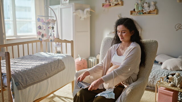 Pregnant woman inspecting baby clothes nursery. Expectant mom holding bodysuit.
