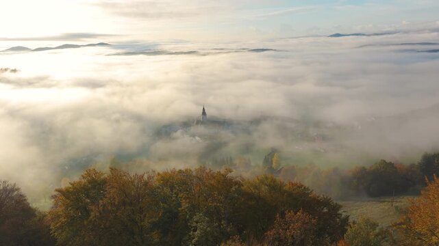 AERIAL: Sticna Abbey emerges softly through thick morning fog in peaceful autumn countryside of Slovenia. Golden trees frame the historic monastery while rolling hills rise above a vast sea of mist.