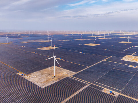 Aerial view of a large-scale solar farm with rows of photovoltaic panels and wind turbines under a cloudy sky in Gujarat, India.