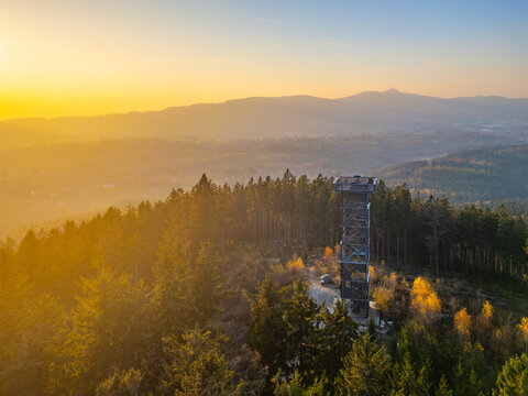 Aerial view of the Cisaruv kamen observation tower rising above a dense coniferous forest during a golden sunset in Liberec 30, Liberec Region, Czechia.