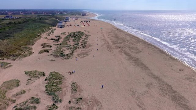 Wide horizontal aerial drone video of Katwijk aan Zee beach resort, North Sea foam waves, sun glint reflections, coastal sand dunes, and town skyline in South Holland, Netherlands on afternoon