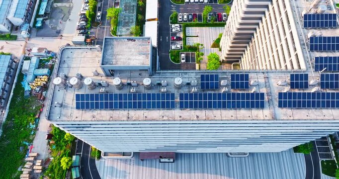 High-angle view of solar panels arranged on an urban rooftop, with metallic storage tanks and adjacent parking lots in bright sunlight.