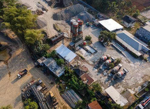 Aerial view of a concrete batching plant featuring tall silos, parked cement mixer trucks, piles of construction materials, and heavy machinery in Cilongok, Central Java, Indonesia.