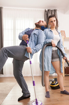 Woman holds a vacuum hose with an unimpressed expression as a man in headphones leans back theatrically with a mop, creating a comic mismatch during shared housework, emphasizing interaction.