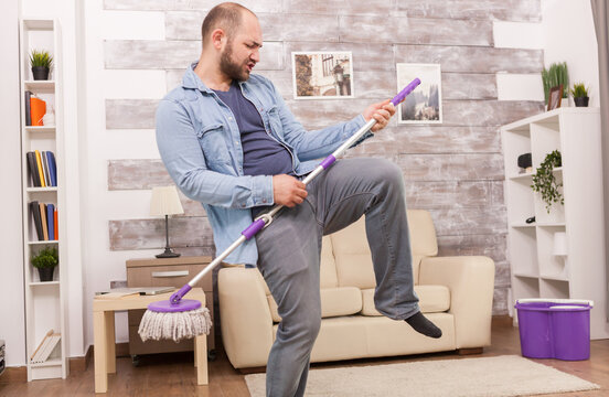 Man holds a mop like a guitar and lifts one knee during housework, creating an energetic and humorous scene about making cleaning more fun.