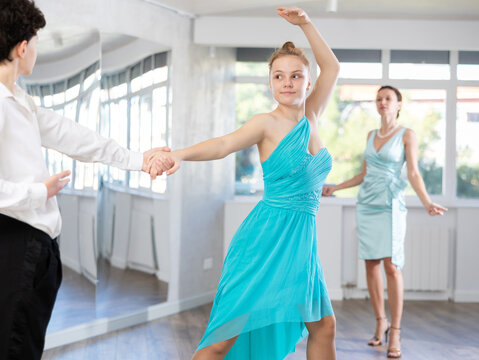 Positive motivated adolescent dance duo, boy and girl, refining slow foxtrot technique in dance hall while interested female coach providing expert guidance