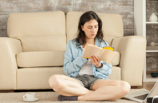 Young woman sits cross-legged on the living room rug reading a book beside an open laptop and coffee cup during relaxed home study, emphasizing relaxation and downtime in everyday home life.