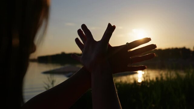 Hands shape a flying bird silhouette backlit by glowing golden hour sun over a lake. Soft ambient flare delivers evocative metaphorical imagery for inspirational storytelling.