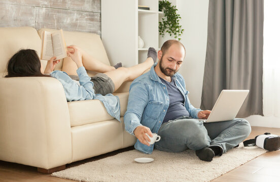 Man reaches for a cup and saucer beside his laptop while a woman reads on the sofa, capturing a casual home break with digital activity and quiet relaxation.
