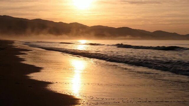Golden hour beach sunset with misty mountains and gentle ocean waves rolling on shore