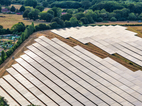 Aerial view of a large solar farm with rows of photovoltaic panels arranged in a field, bordered by lush green trees and rural landscape in Hook, England, United Kingdom.
