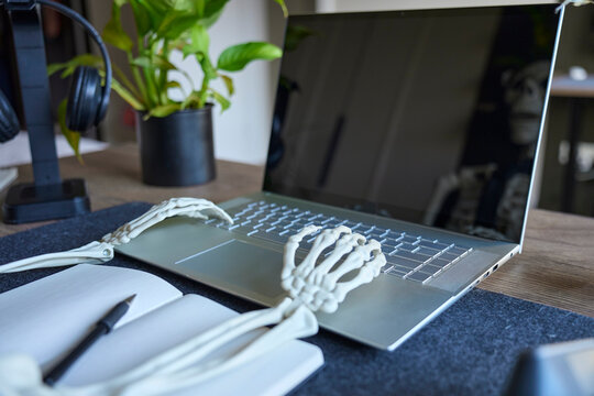 Skeleton hands typing on laptop keyboard beside notebook on desk