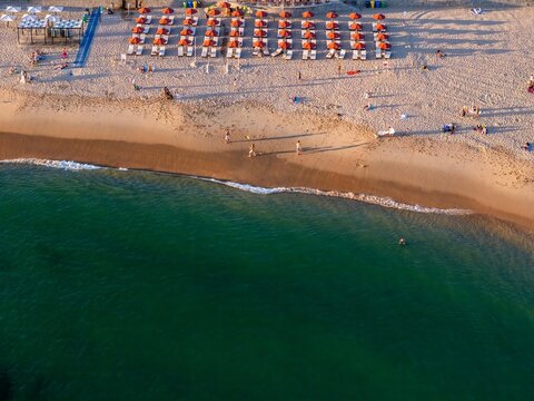 Aerial view of Tamariz Beach with rows of orange umbrellas on the sand, clear turquoise water, and people walking along the shoreline in Estoril, Lisbon, Portugal.