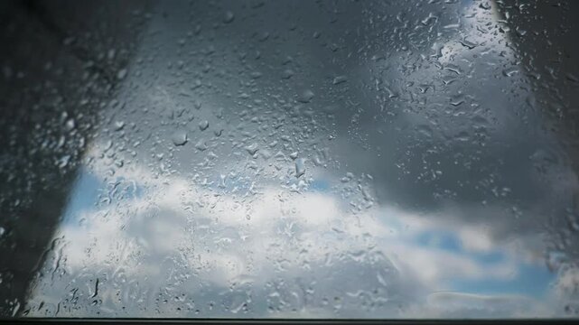 Raindrops cling and slide down a clear glass pane against a cloudy sky. The moody ambient lighting provides versatile atmospheric texture for reflective narrative sequences.