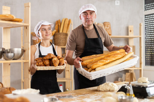 Home production of bread, work room. Grandpa and teen schoolgirl standing in kitchen with basket of finished products baguettes, croissants. Girl participates in family business, baking bread products