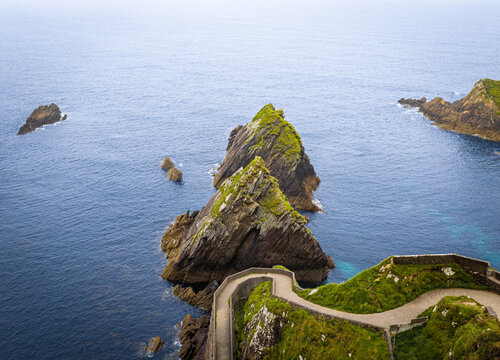 Aerial view of Dunquin Pier with its winding stone path leading down to the rugged coastline, surrounded by dramatic rocky cliffs and the deep blue Atlantic Ocean in County Kerry, Ireland.