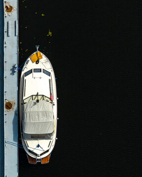 Aerial view of a white motor boat with a wooden deck and grey canvas cover moored at a concrete pier in the dark water of a port in Poland.