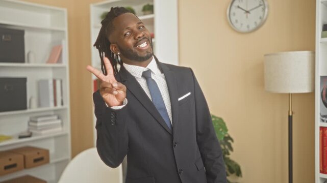 Man in dark suit smiling and flashing a two finger peace sign with his hand in an office building near bookshelf, clock and floor lamp; confidence.