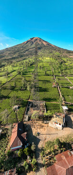 Aerial view of Sikatok Cell Tower standing amidst lush green tea plantations and rural houses with a majestic mountain in the background in Kecamatan Kejajar, Jawa Tengah, Indonesia.