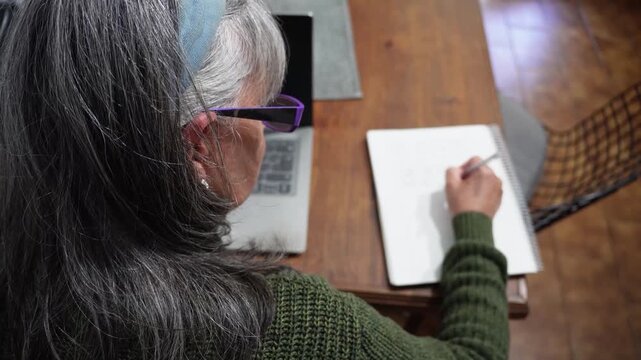 Elderly woman writing in notepad during online study session
